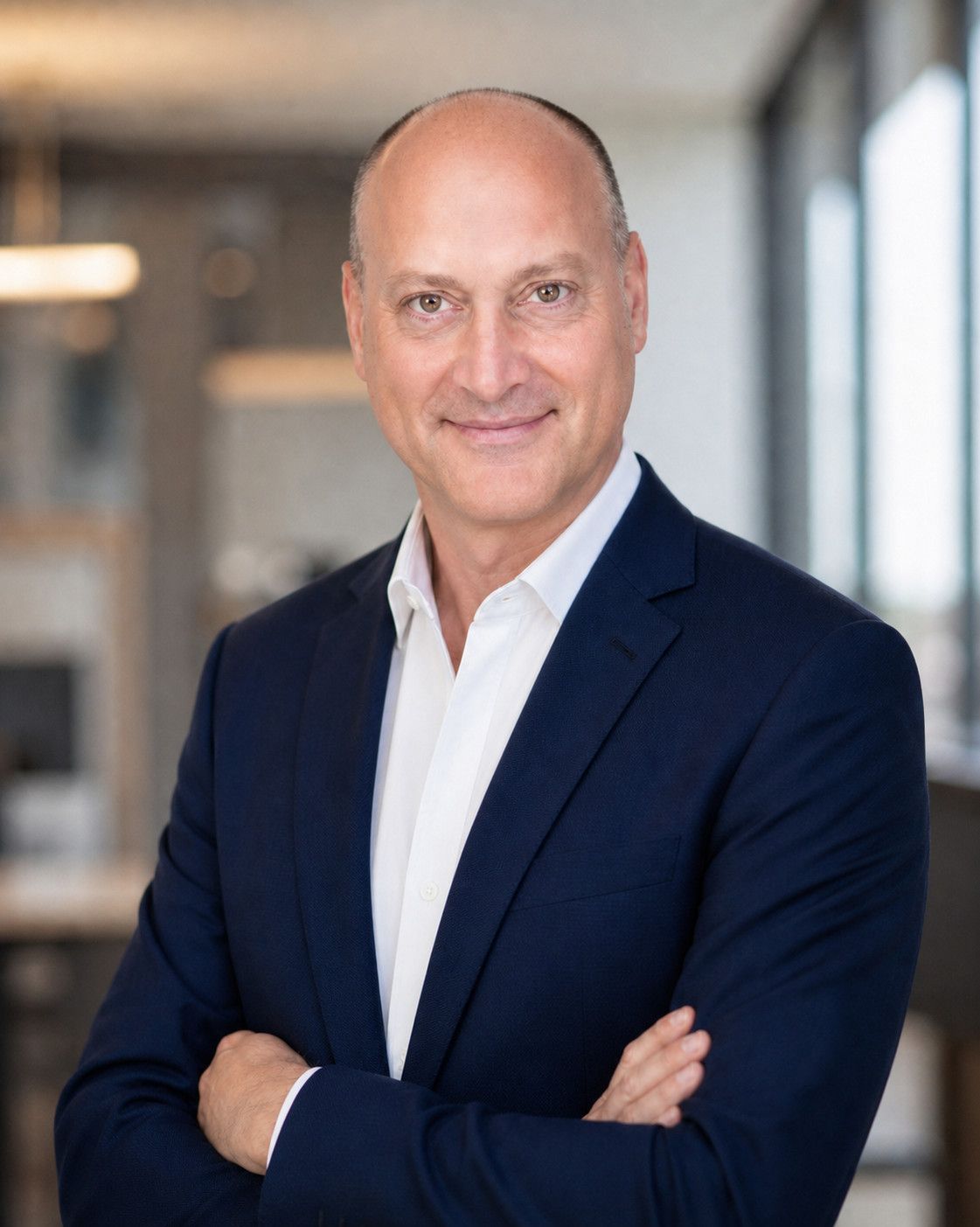 Peter DeCaprio standing with folded arms in a bright office, in a navy suit and white open-collared shirt